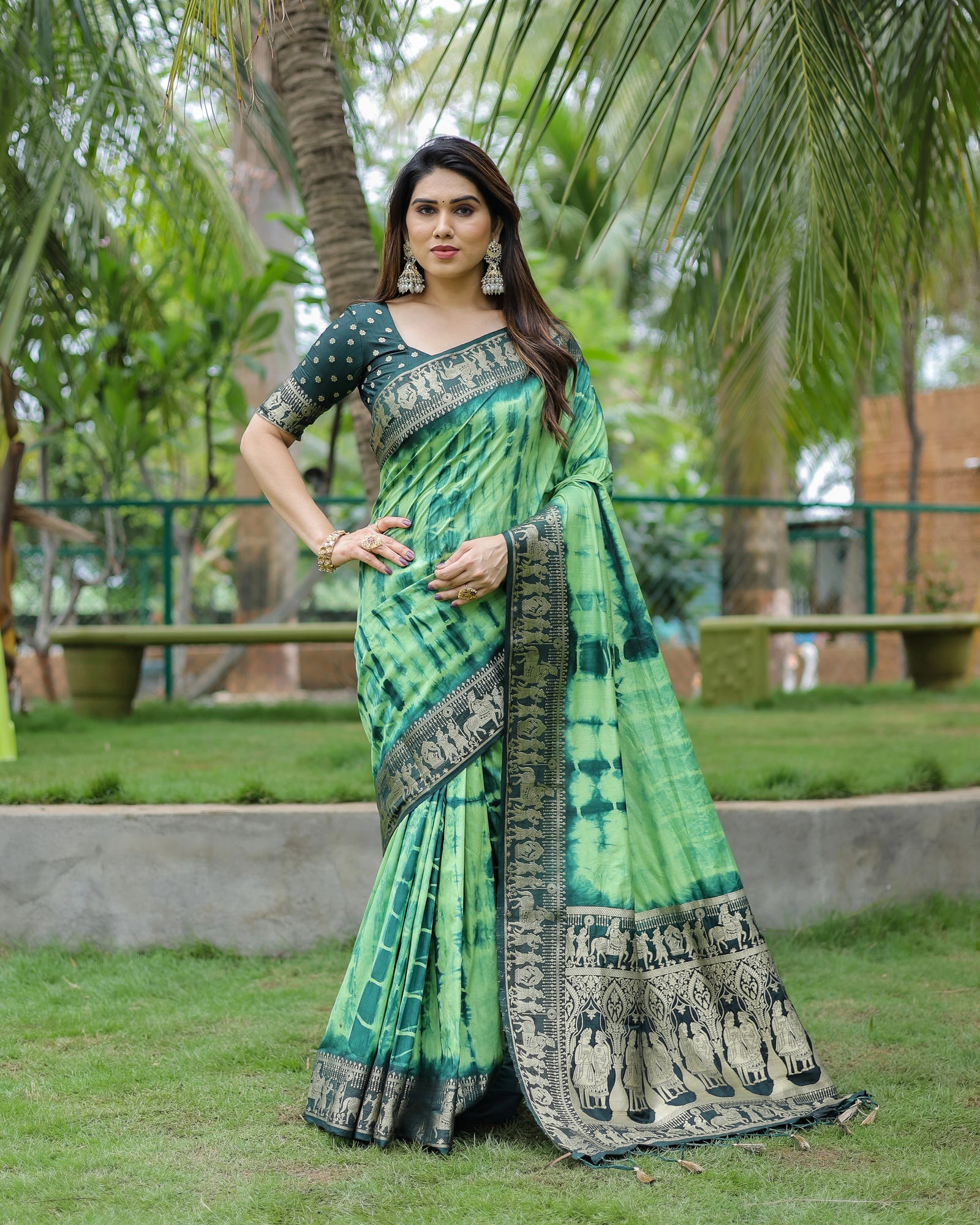 Woman in a green saree with black patterns standing outdoors with trees in the background