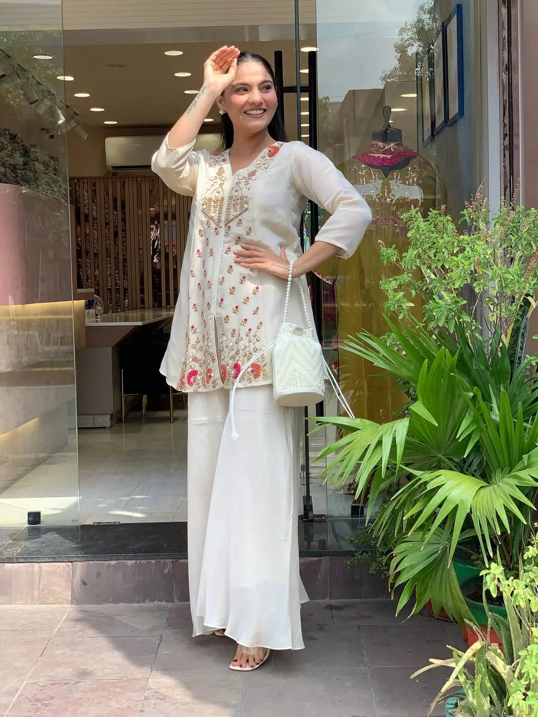 Woman in a white traditional outfit standing outside a building with plants in the foreground.