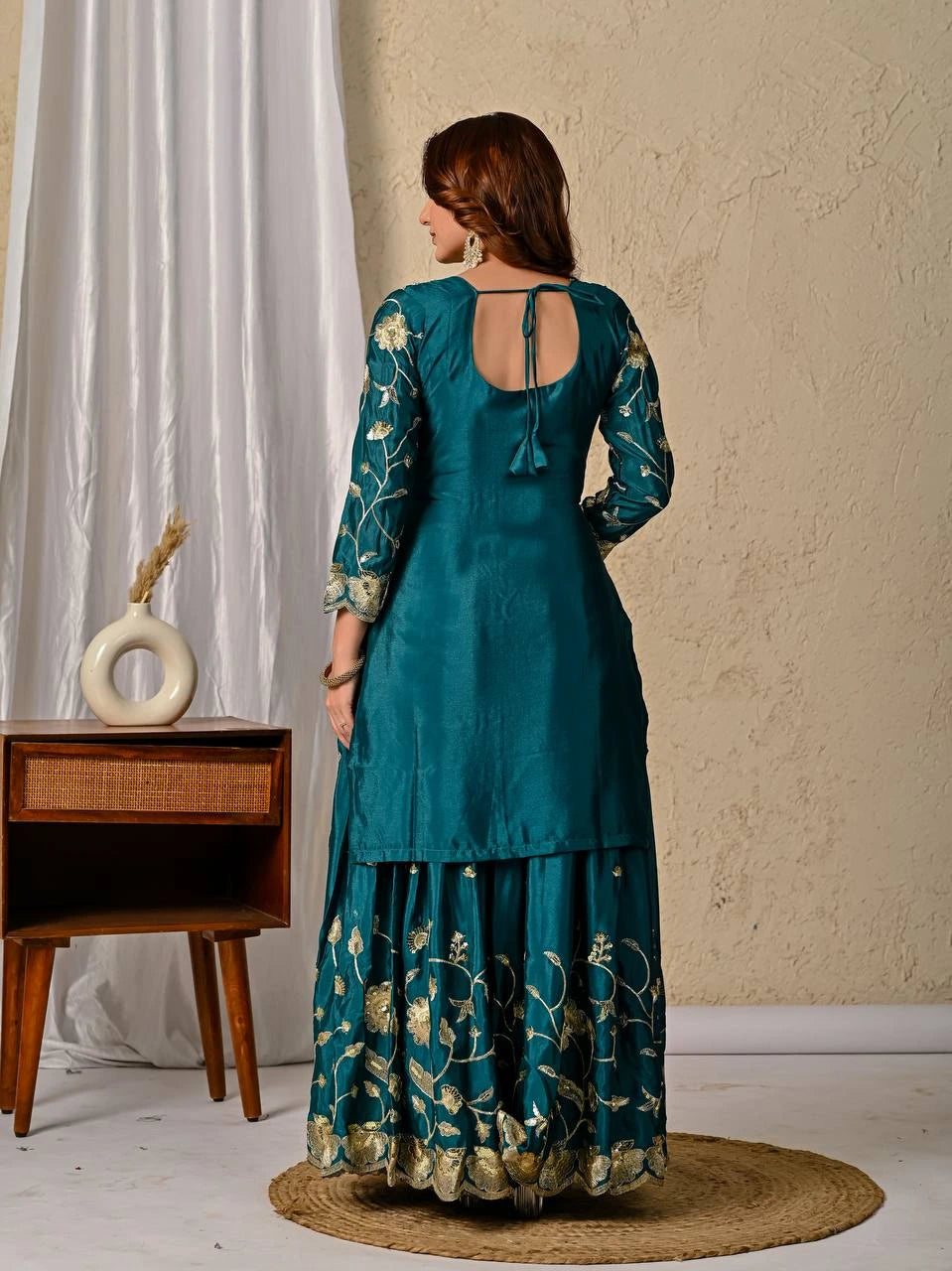 Woman wearing a teal traditional outfit with floral patterns in a minimalistic room.