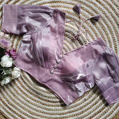 Lavender satin blouse with pleated detailing and four decorative silver buttons on the front, featuring elbow-length sleeves and tie-up tassels, displayed on a circular woven mat with white and purple flowers on the side