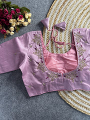 A lavender-toned raw silk blouse featuring a deep, square-rounded back neckline. The edges are decorated with hand-embroidered floral sprigs in gold and pearl, accented with deep magenta threadwork. It includes matching lavender tie-back cords with triangular tassels, and small pearl floral buttis are scattered across the back and sleeves.