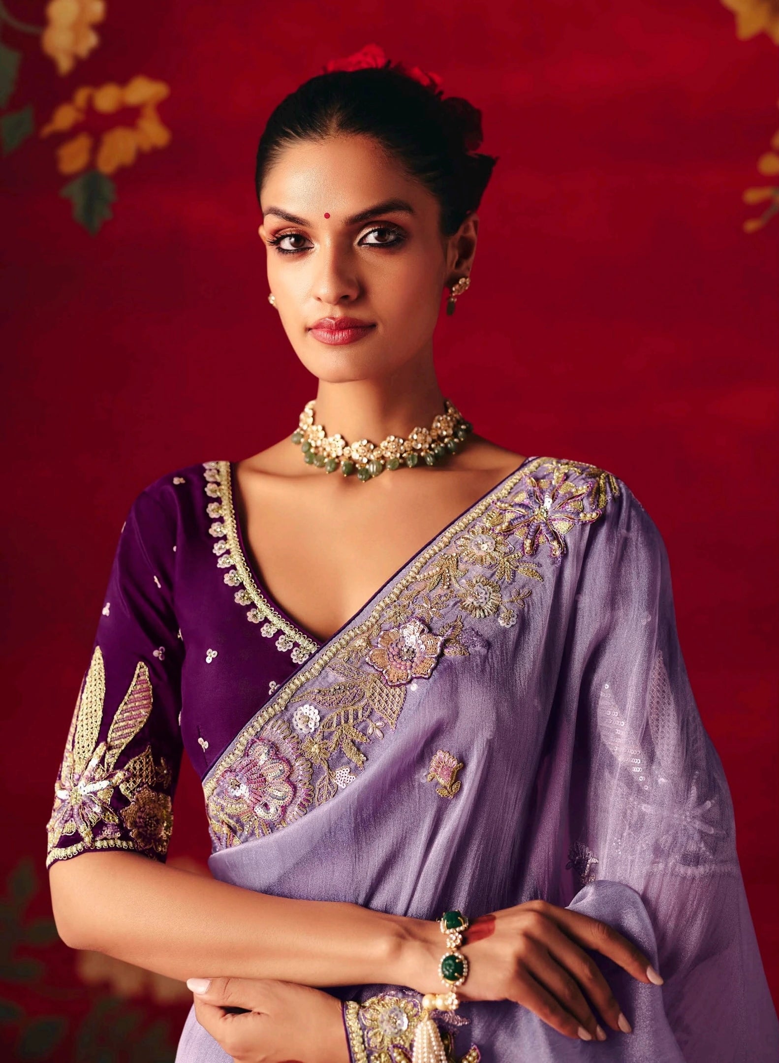 Woman wearing a purple saree with gold embroidery against a red background