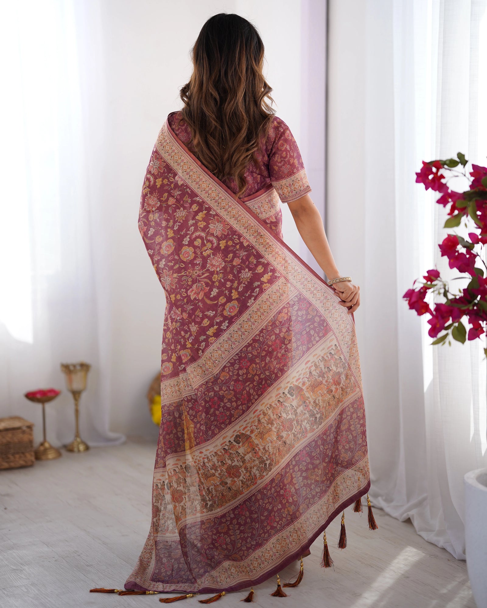 Woman wearing a patterned saree in a bright room with white curtains and flowers.