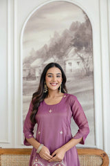Woman in a purple outfit standing in front of a decorative wall with a landscape painting.