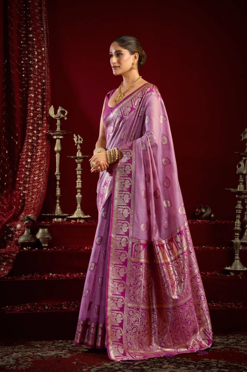 Woman in a pink saree with intricate patterns standing against a red curtain backdrop.