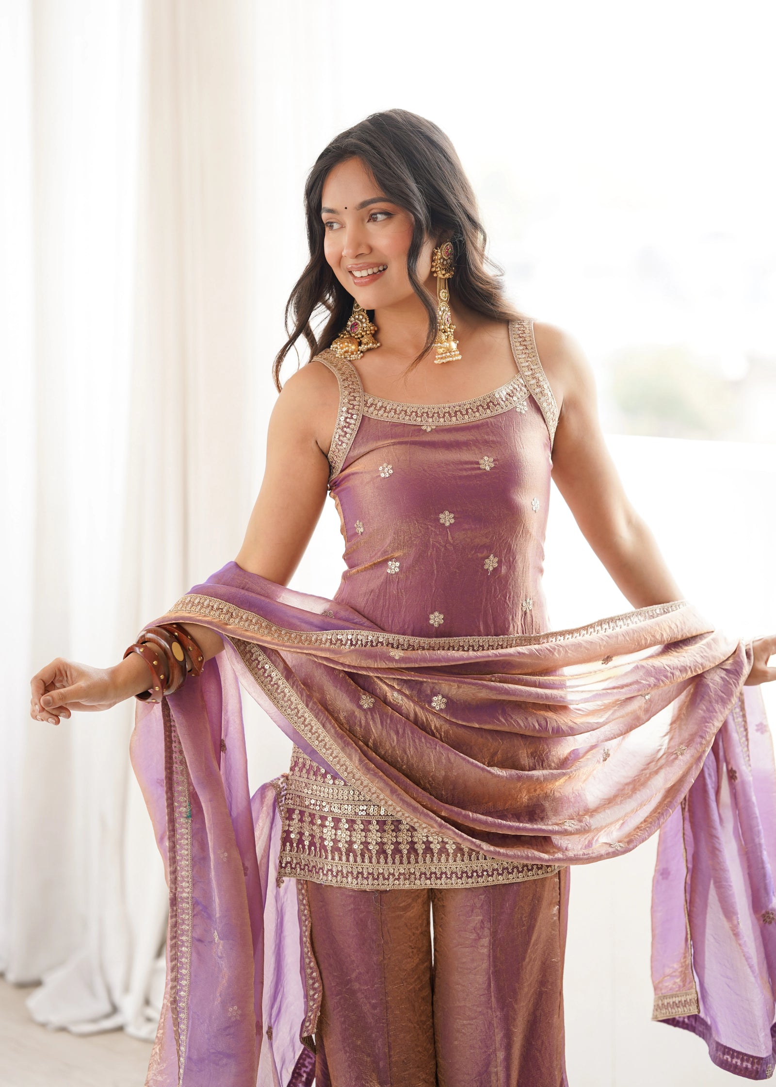 Woman in a traditional outfit with a purple dupatta, standing indoors.