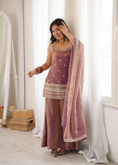 Woman in a traditional outfit with a purple top and brown pants, standing in a room with white curtains.