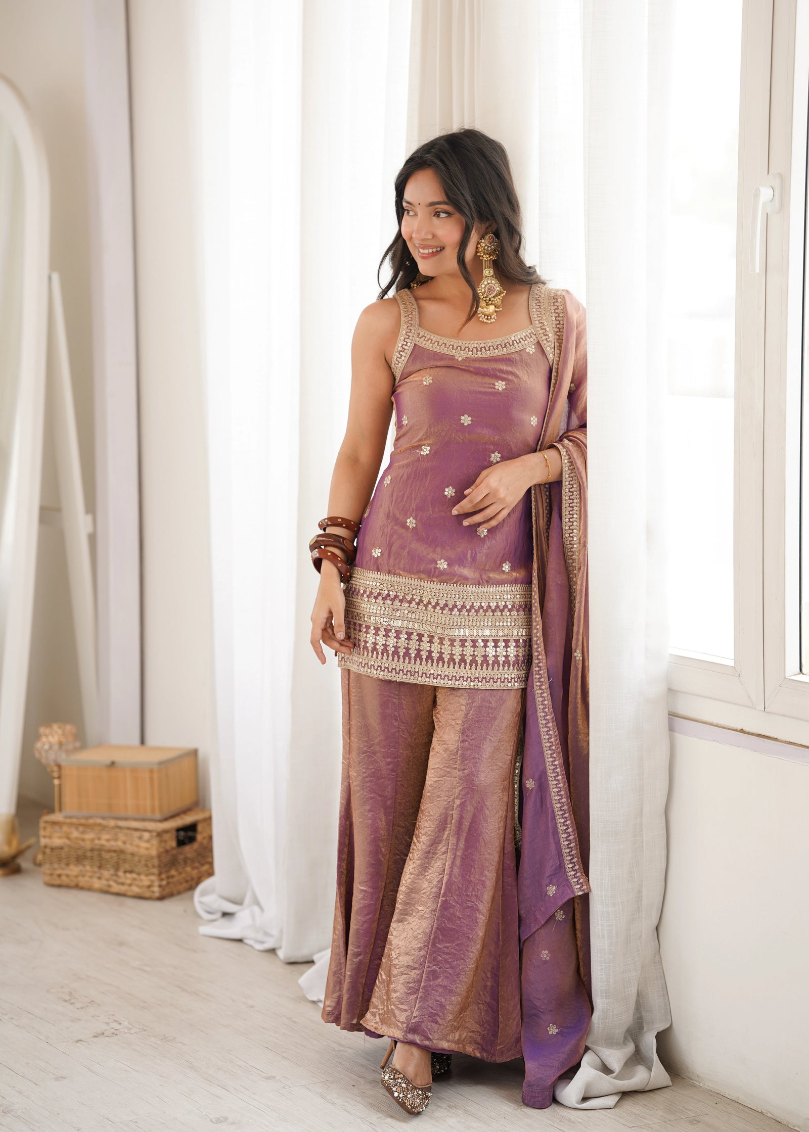 Woman in a traditional outfit standing in a room with white curtains and wooden boxes.