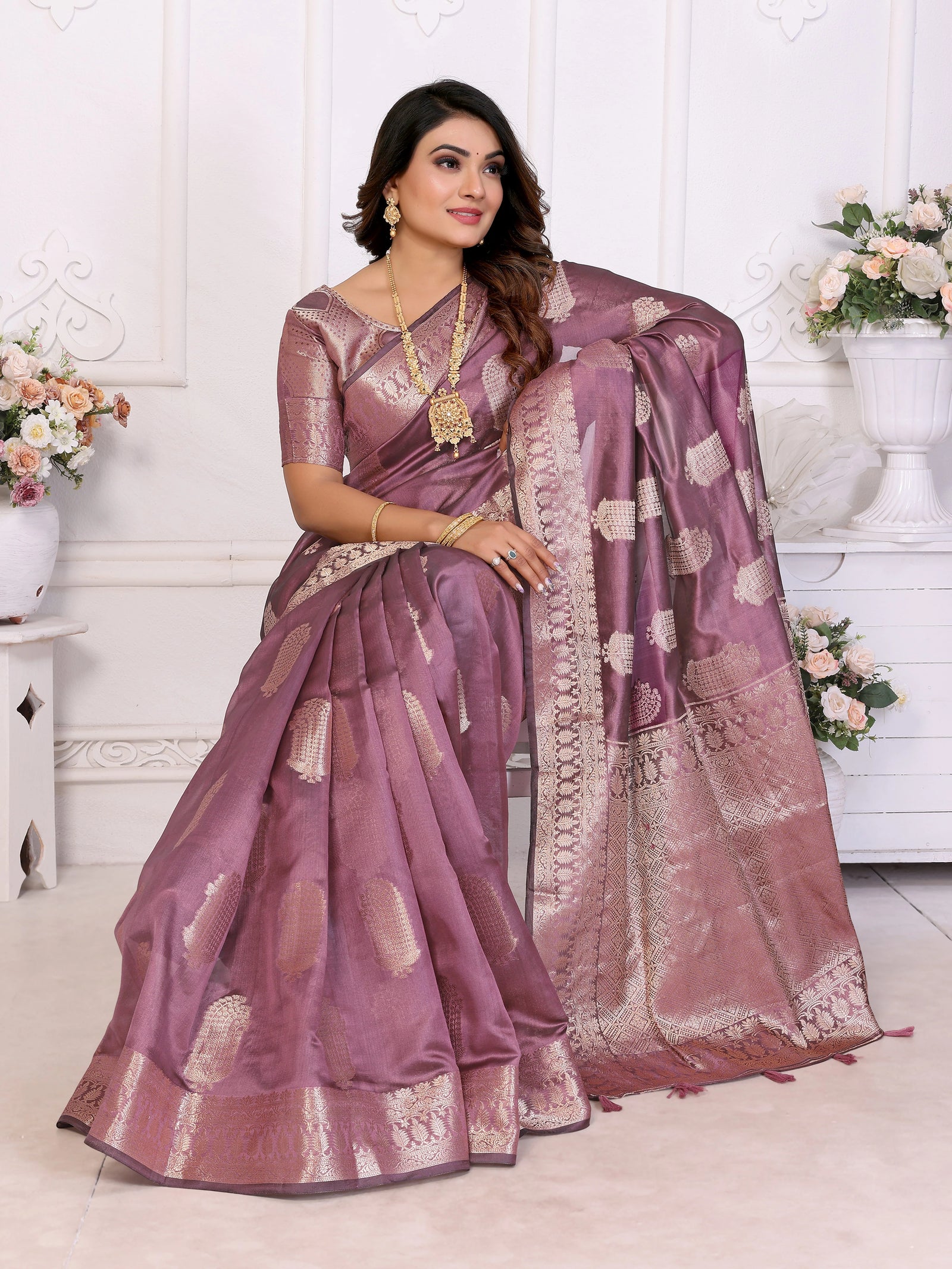 Woman wearing a purple saree with gold patterns in a decorated room.
