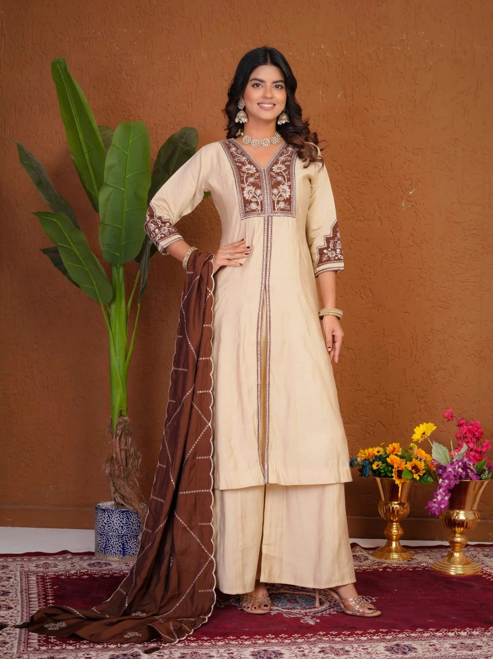 Woman in a traditional outfit with embroidery, standing against a brown wall with decorative elements.