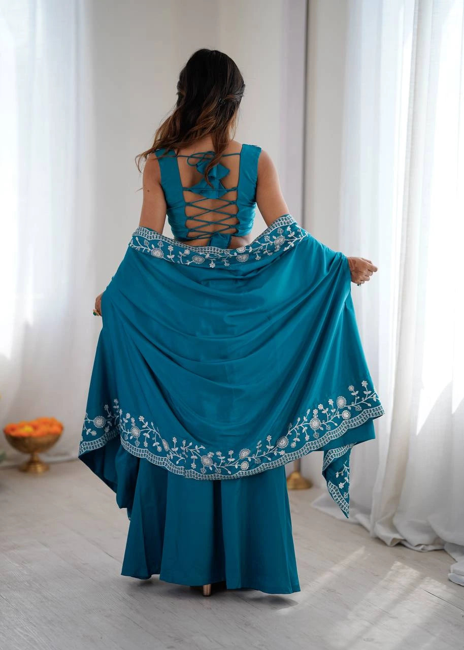 Woman wearing a teal traditional outfit with intricate designs, standing in a room with white curtains.