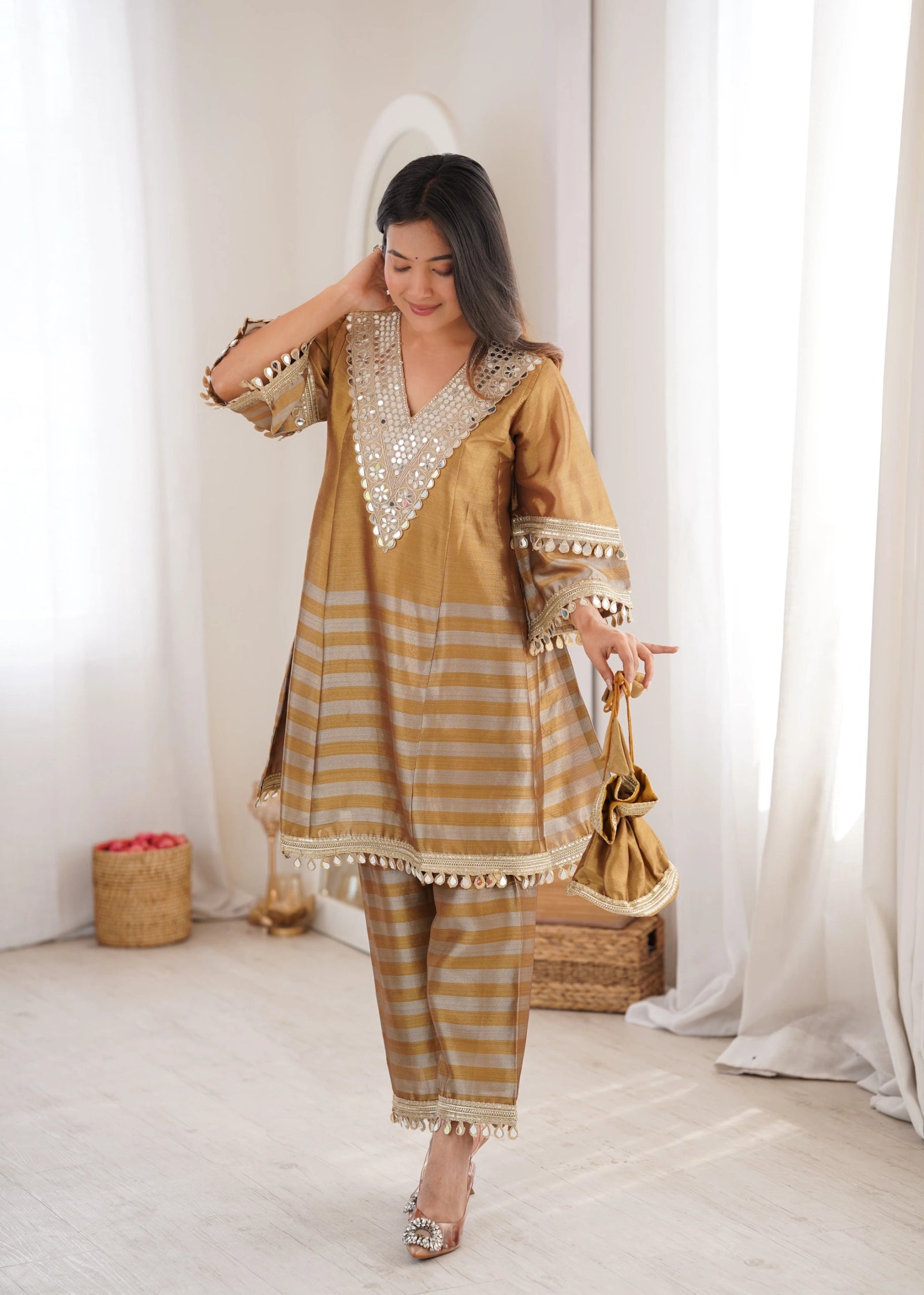 Woman in a gold and white traditional outfit holding a handbag in a bright room.