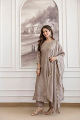 Woman in a beige traditional outfit standing in a room with white walls and an arched window.