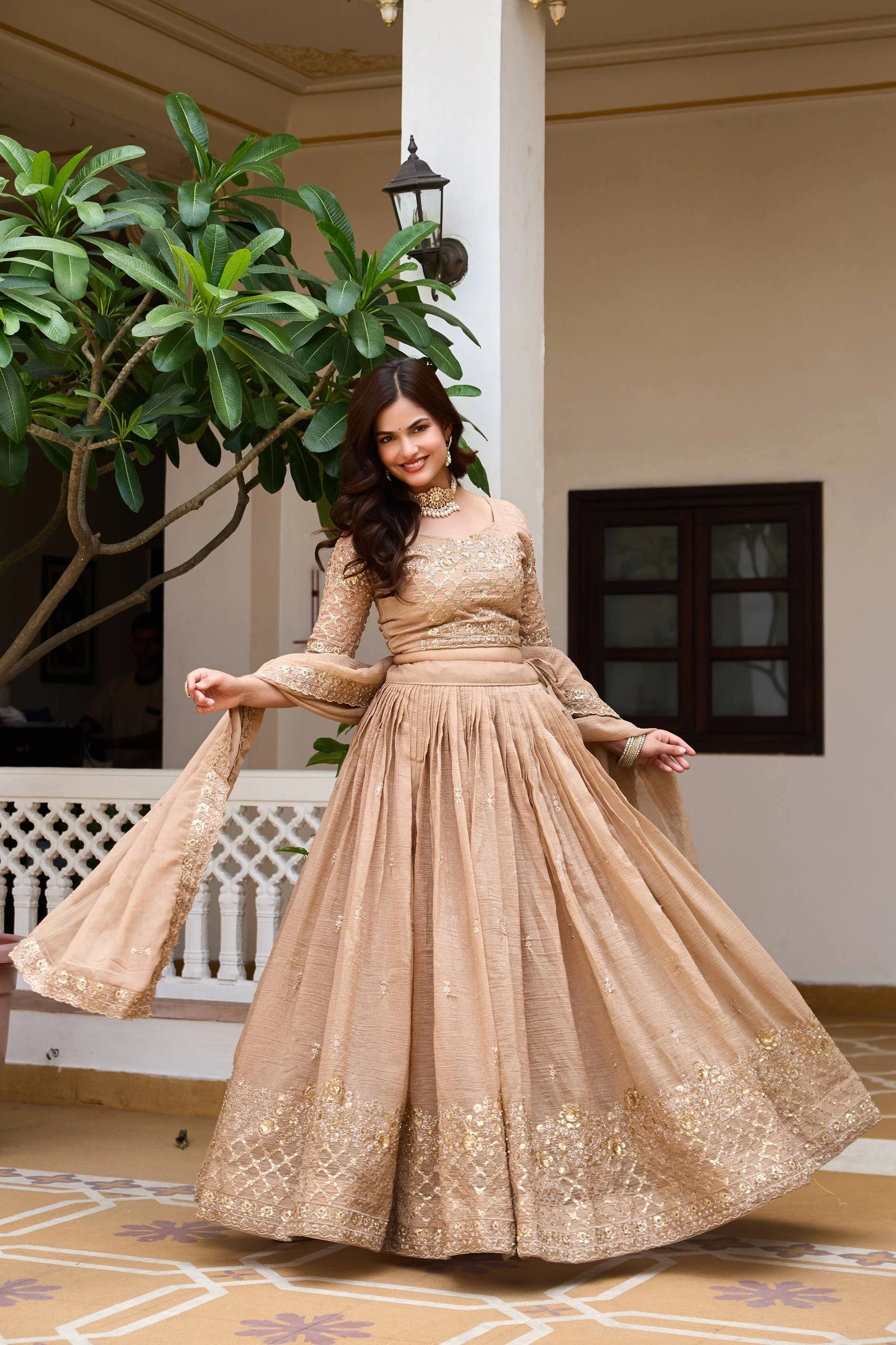 Woman in a beige embroidered dress standing in a decorative indoor setting with plants and architectural elements.