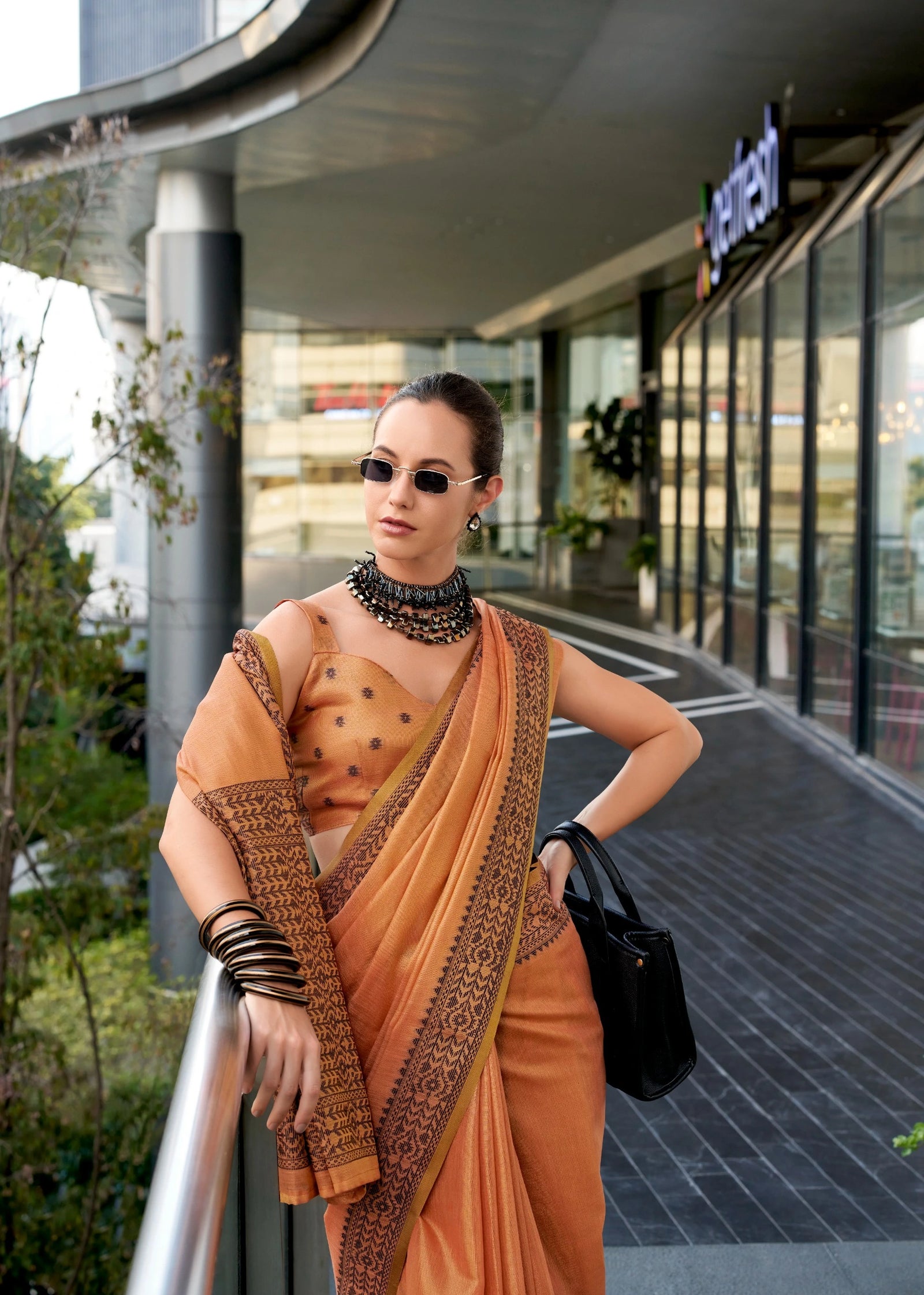 Woman in an orange saree with a patterned blouse standing outdoors near a building.