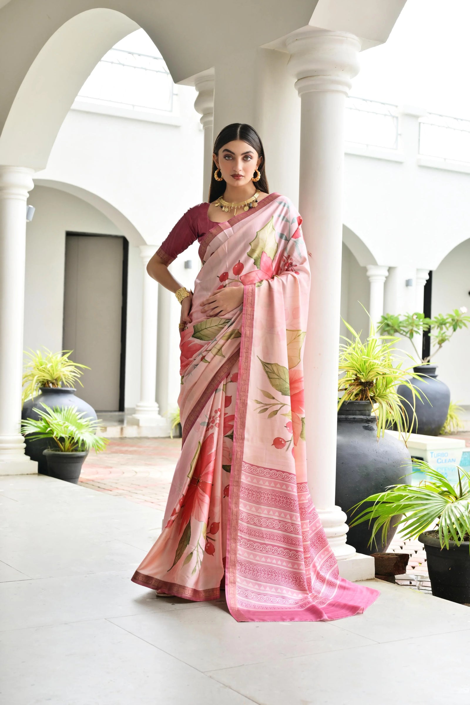 Woman in a pink saree with floral patterns standing in an elegant outdoor setting.