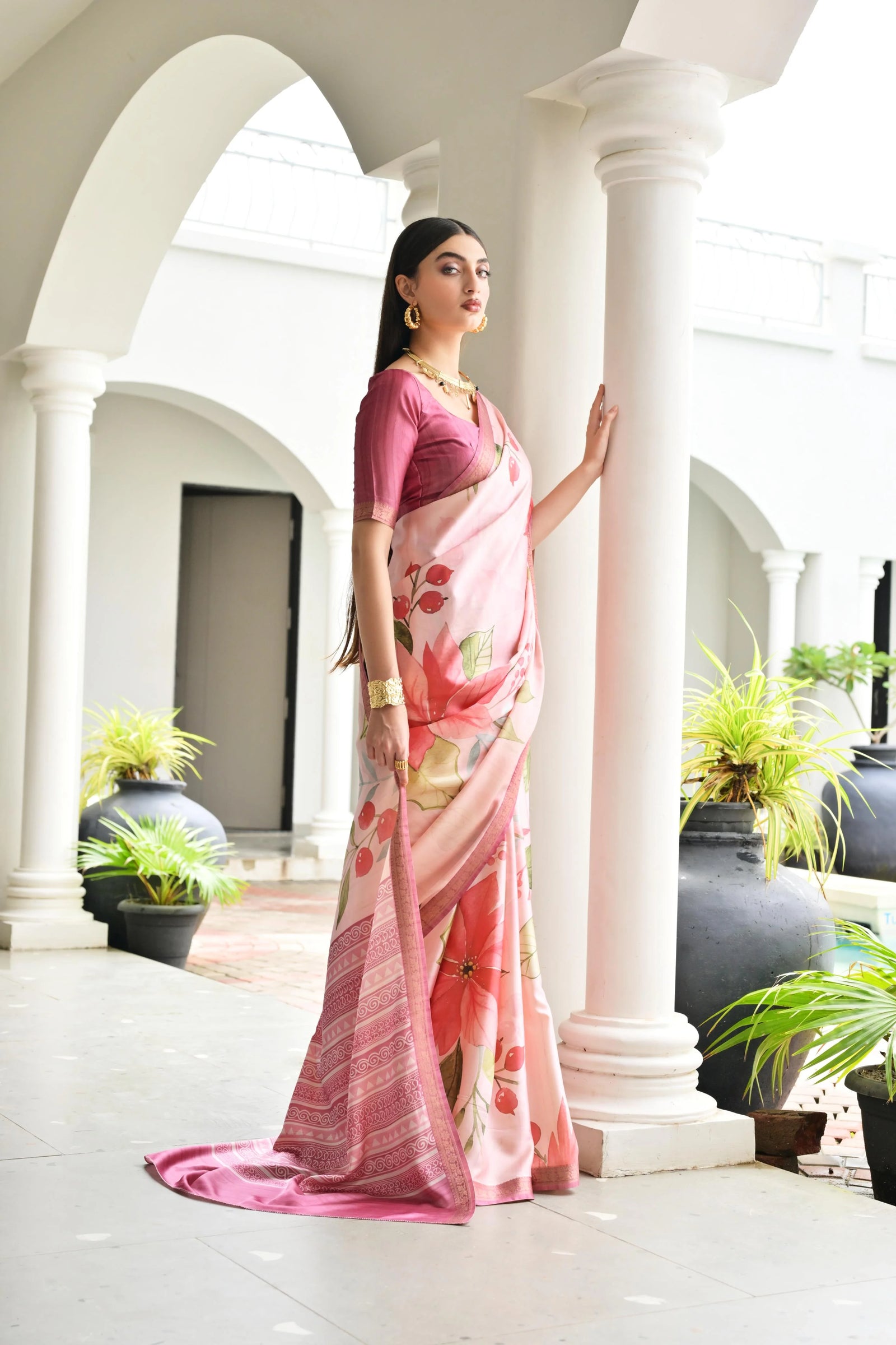 Woman in a pink saree with floral patterns standing in a well-lit indoor setting with columns and plants.