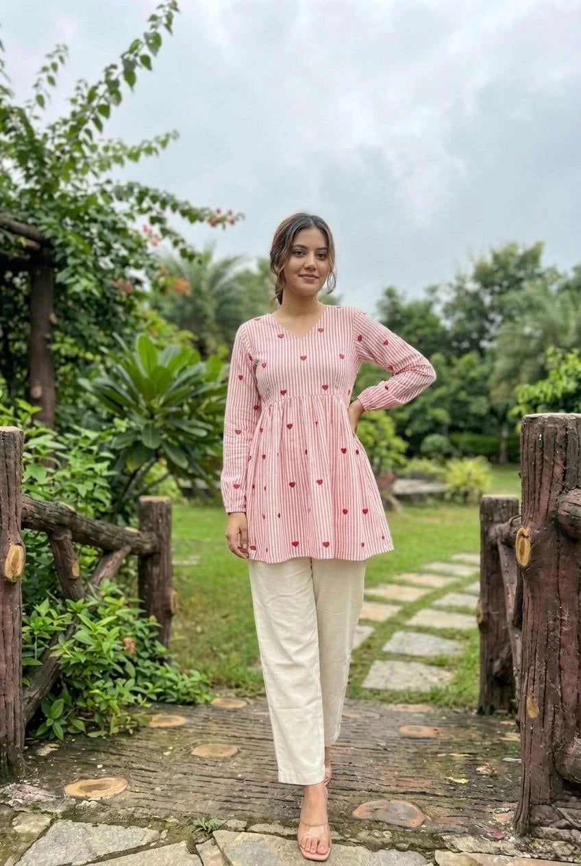 Woman in a pink checkered top and white pants standing on a wooden bridge in a garden.