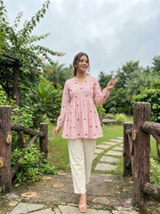 Woman in a pink top and white pants standing in a garden with wooden fences and greenery.