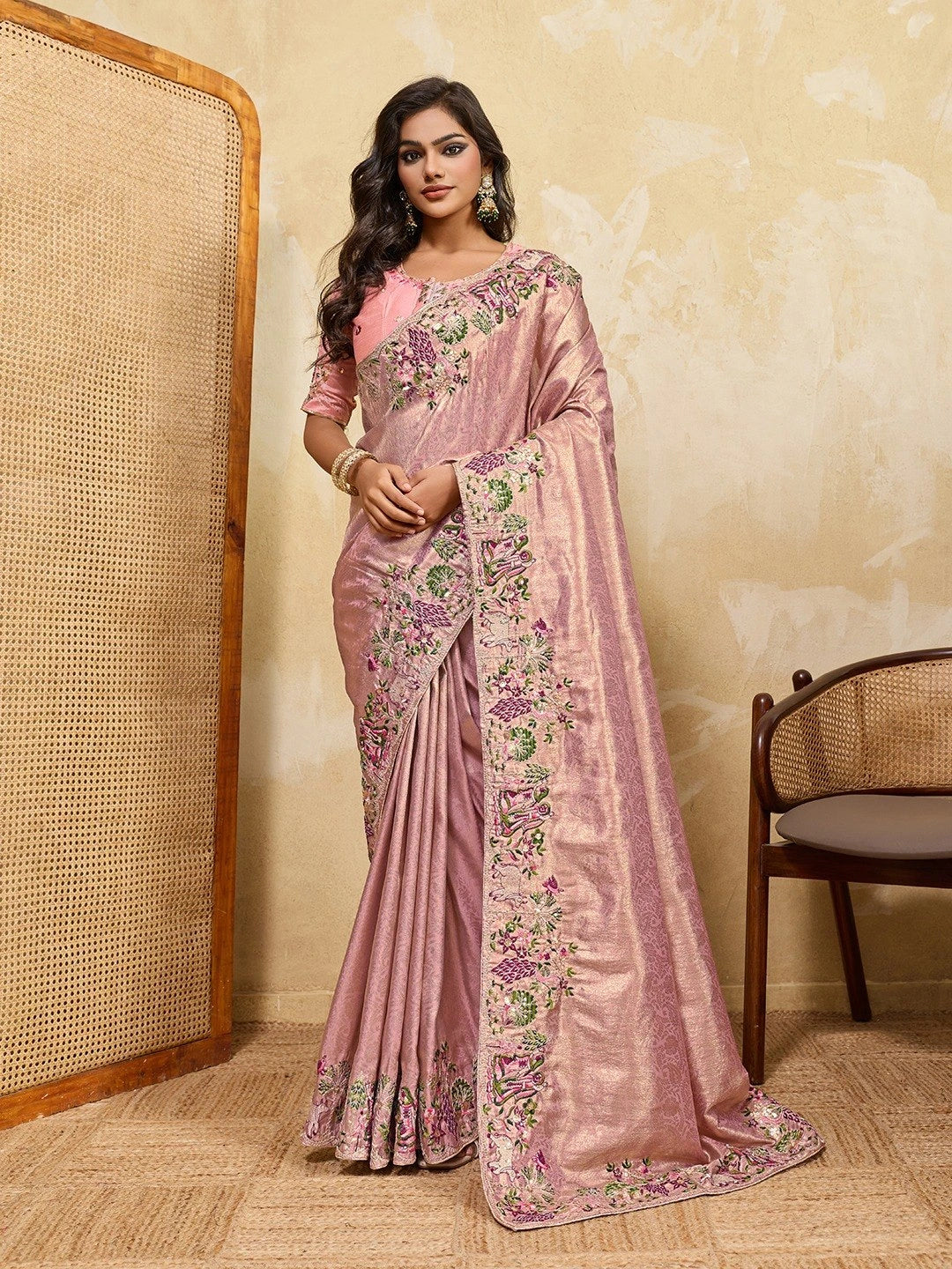 Woman wearing a pink saree with floral patterns in a room with a textured wall and chair.