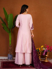 Woman in a light pink traditional outfit with a purple dupatta standing against a brown wall with decorative elements.