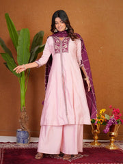 Woman in a pink traditional outfit with purple accents standing against a brown wall with decorative elements.