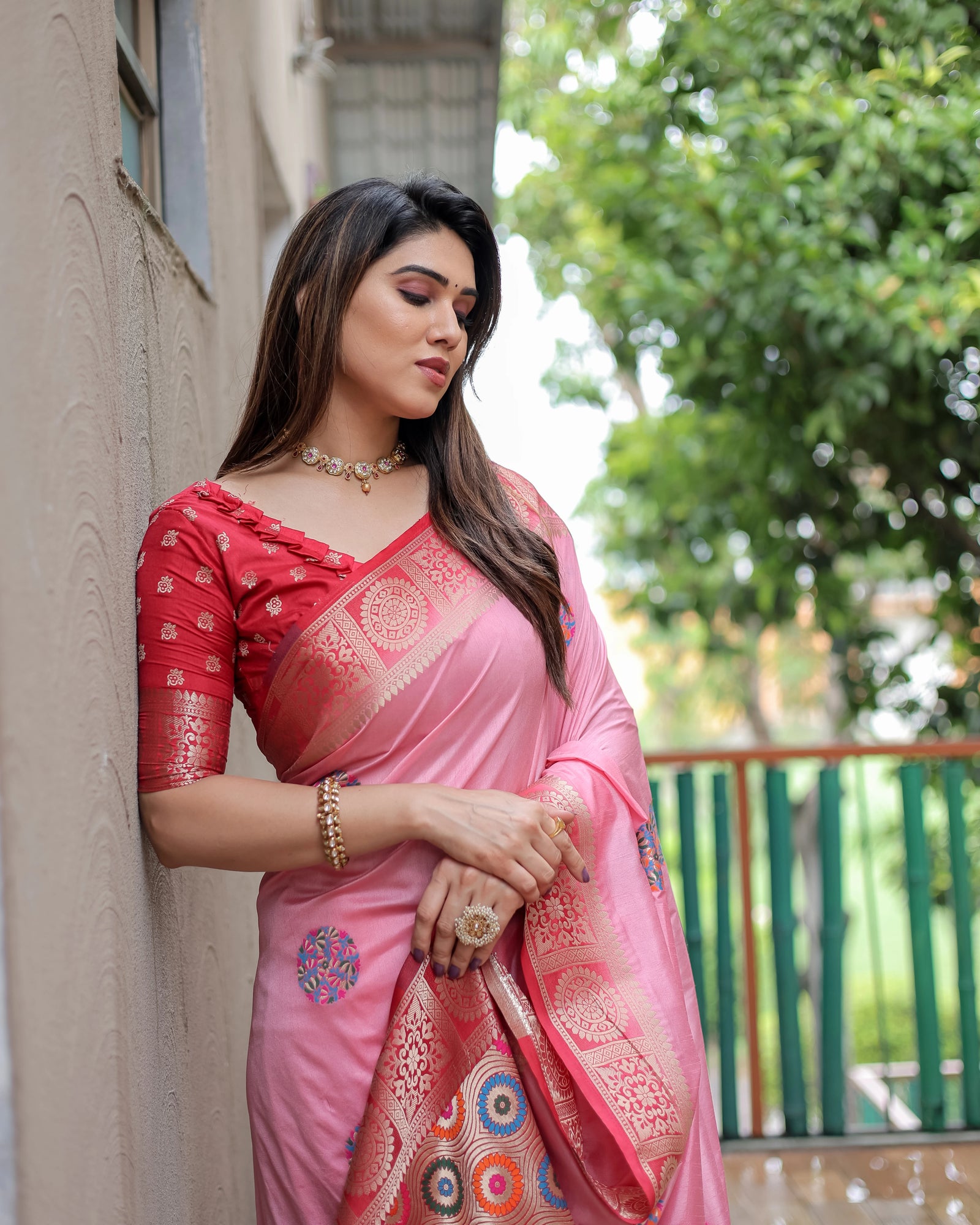 Woman in a pink saree leaning against a wall with greenery in the background