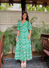 Woman in a green floral dress standing in an outdoor setting with plants and a textured wall.