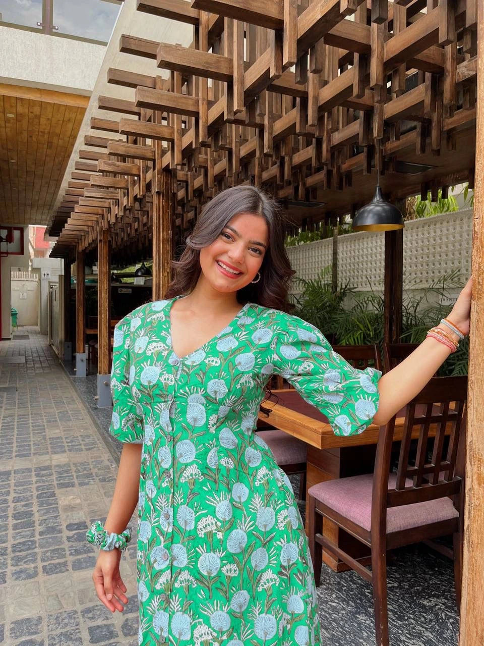 Woman in a green dress standing outdoors under a wooden architectural structure.