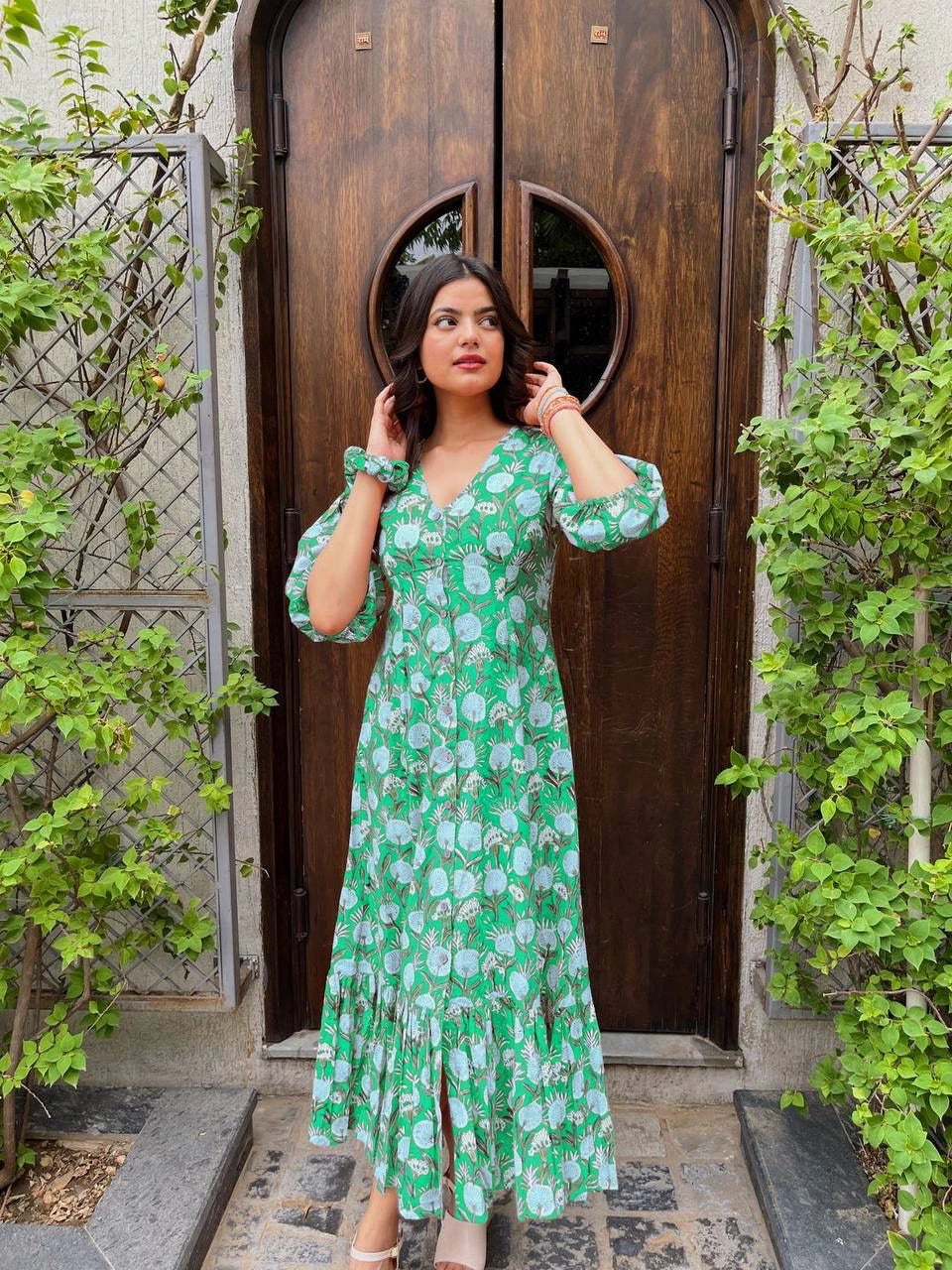 Woman in a green floral dress standing in front of a wooden door with plants around.
