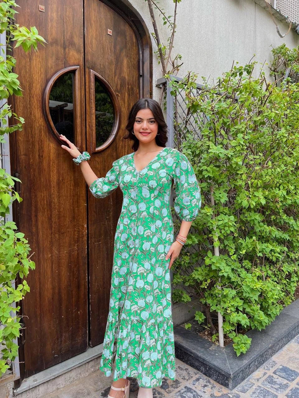 Woman in a green floral dress standing next to a wooden door with circular design.