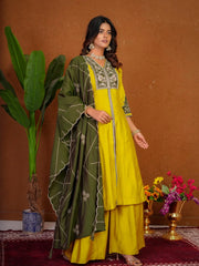 Woman wearing a yellow and green traditional outfit with jewelry, standing against a brown wall.