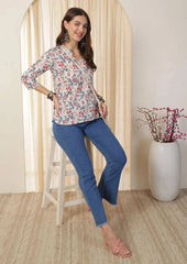 Woman in floral blouse and blue jeans posing in a room with a decorative plant.
