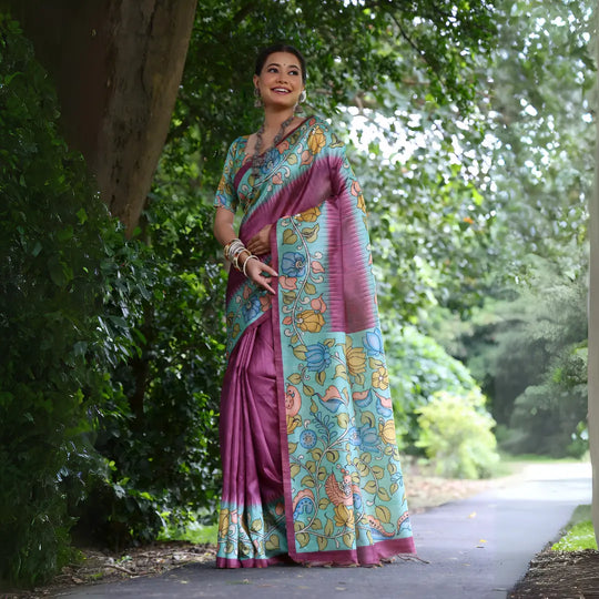 A woman models a stunning Magenta Soft Tussar Silk Saree. The pallu features a wide Sky Blue panel with an elaborate Kalamkari-style print of pink, yellow, and blue floral/peacock motifs. She wears a matching short-sleeve printed blouse and accessorizes with heavy oxidized silver jewelry, posing outdoors in a garden.