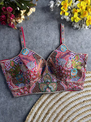 Traditional multicolor bustier blouse featuring vibrant Madhubani-inspired embroidery with intricate floral, paisley, and geometric motifs, embellished with sequins and beads, structured cups, and adjustable straps, displayed flat on a textured grey surface with artificial pink, yellow, and white flowers in the corners.