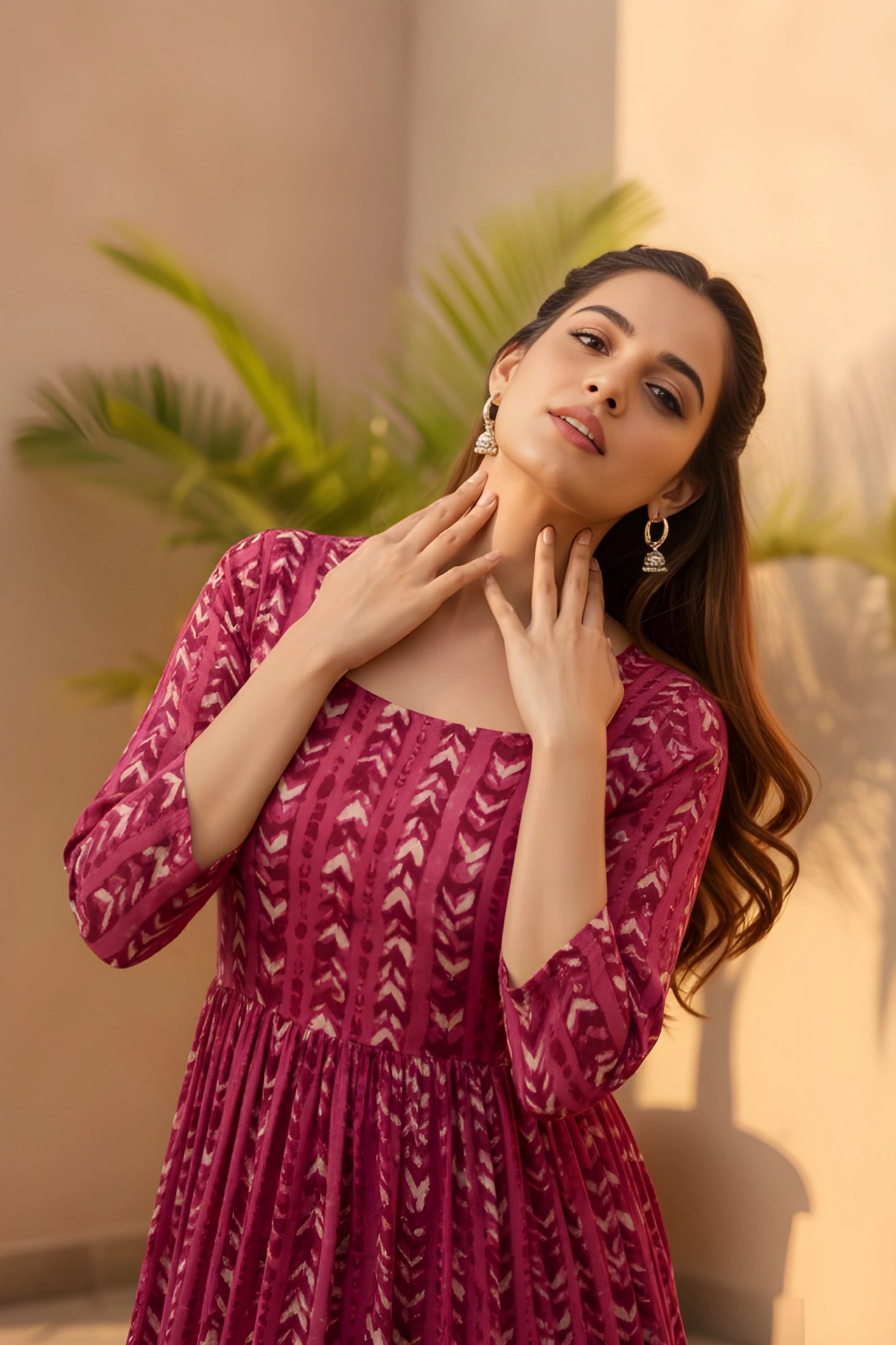 Woman in a pink patterned dress posing with hands on neck against a neutral background