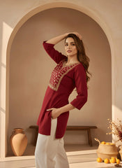 Woman wearing a red embroidered top and white pants in a warm-toned interior setting.