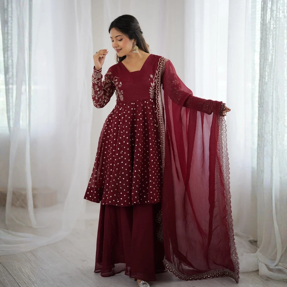 Woman in a maroon traditional outfit with a dupatta against a white curtain background