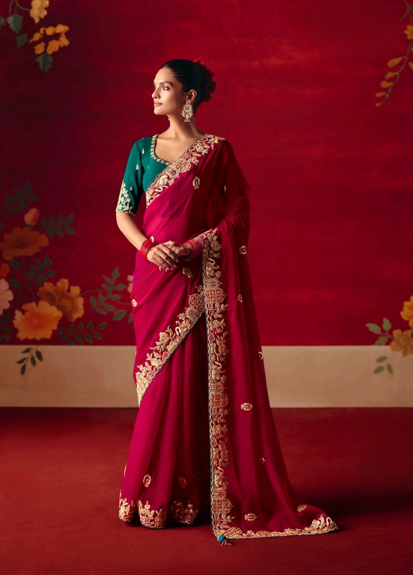 Woman in a red saree with gold border against a floral backdrop