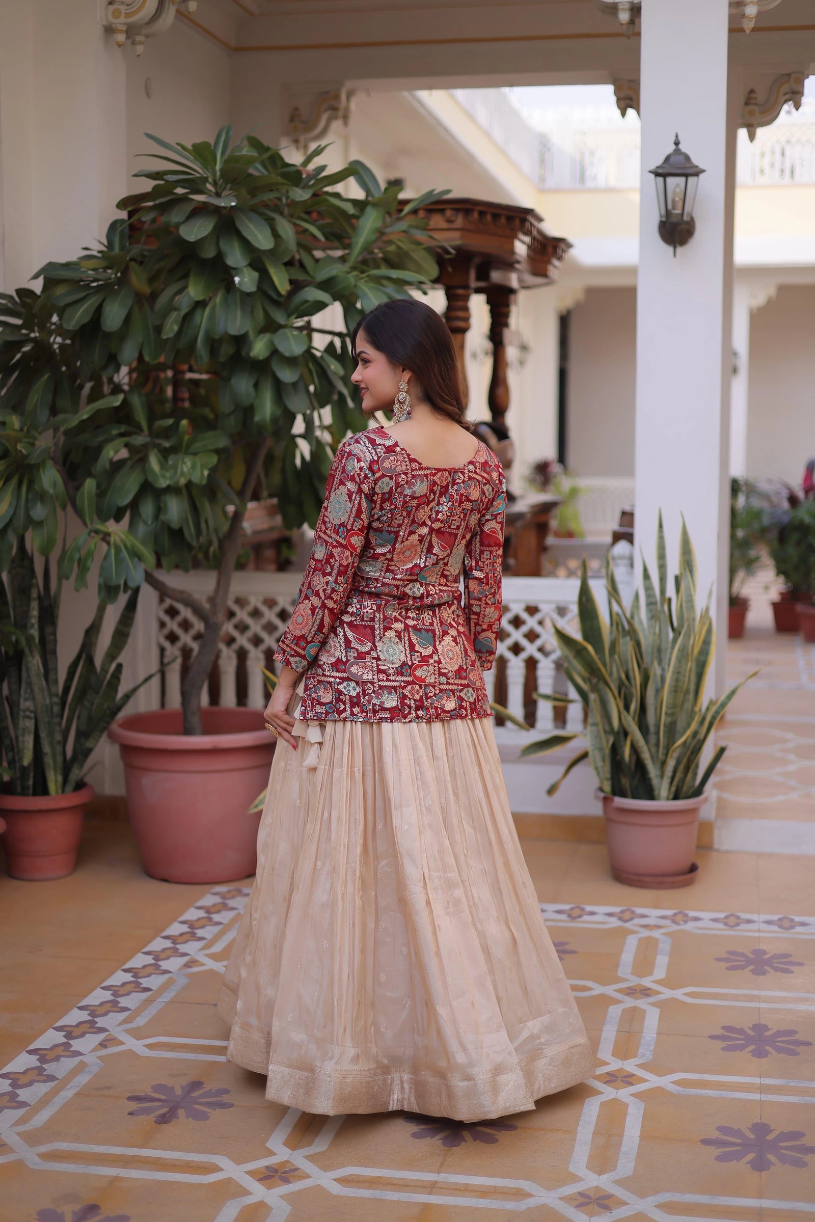 Woman in traditional outfit standing in a decorated indoor setting with plants and patterned floor.