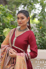 Woman in traditional attire with a red blouse and patterned dupatta, standing outdoors with greenery.