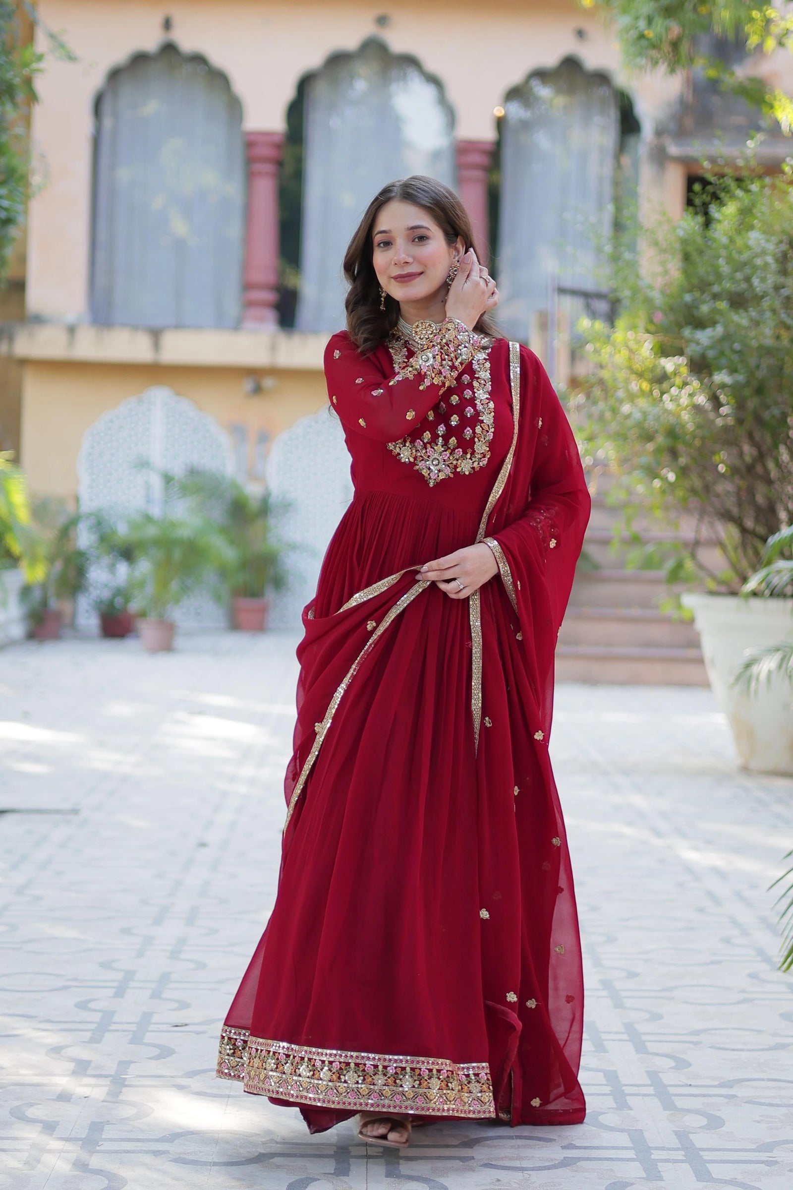 Woman in a red saree with gold embroidery standing outdoors.