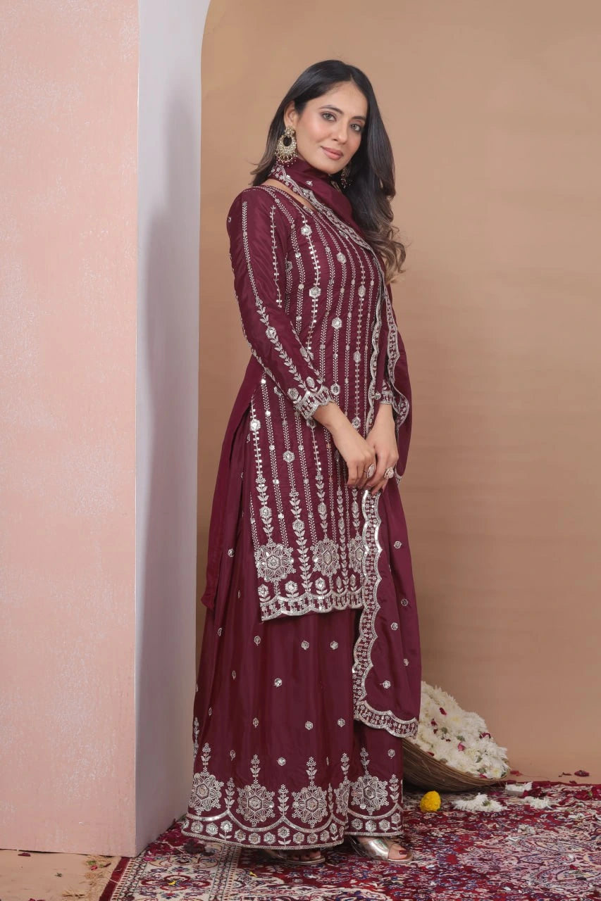 Woman in a maroon embroidered traditional outfit standing against a beige wall.