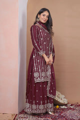 Woman in a maroon embroidered traditional outfit standing against a beige wall.