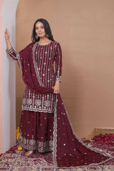 Woman wearing a maroon traditional outfit with white embroidery against a beige wall.