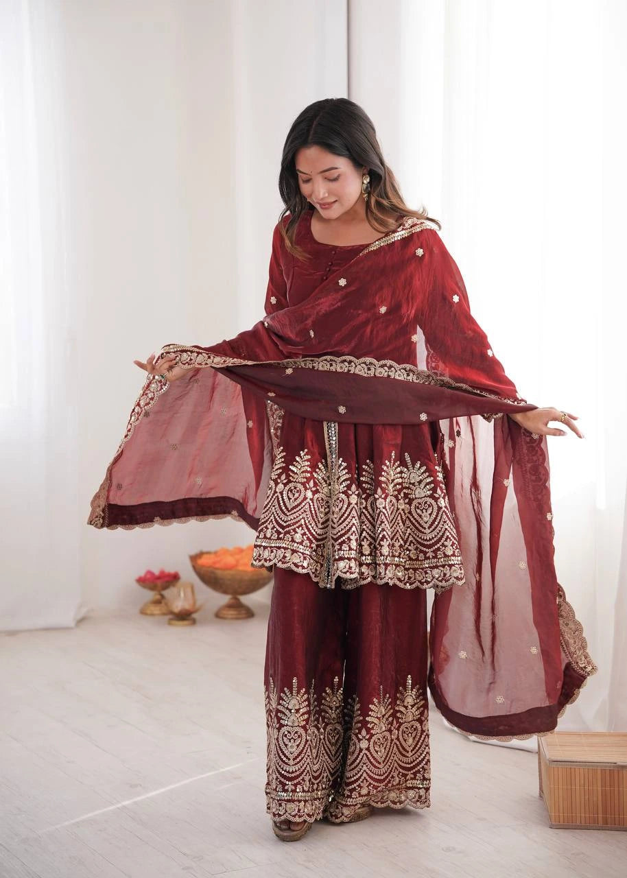 Woman wearing a traditional maroon outfit with gold embroidery in a minimalistic setting.