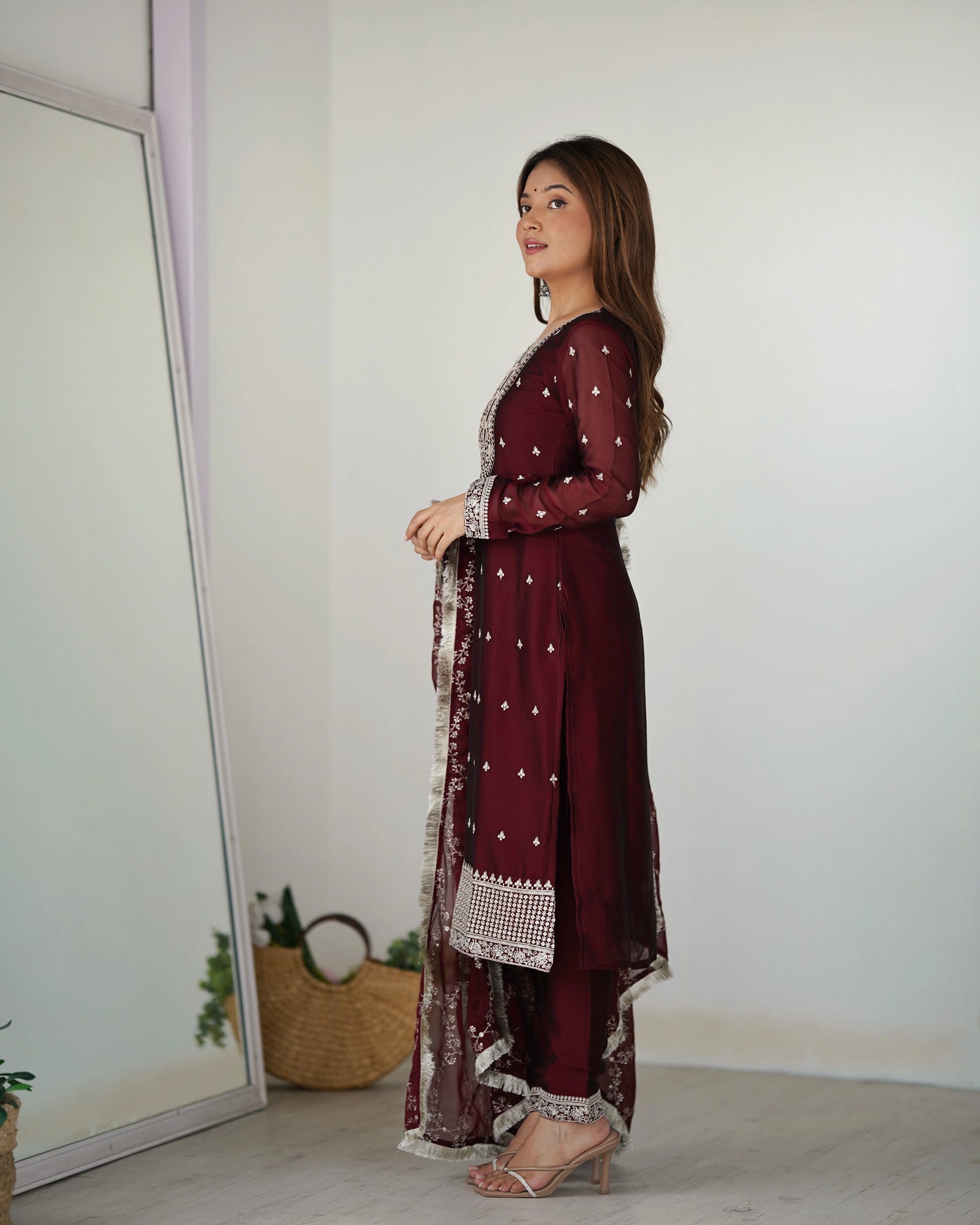 Woman in a burgundy traditional outfit standing in a room with a mirror and plants.
