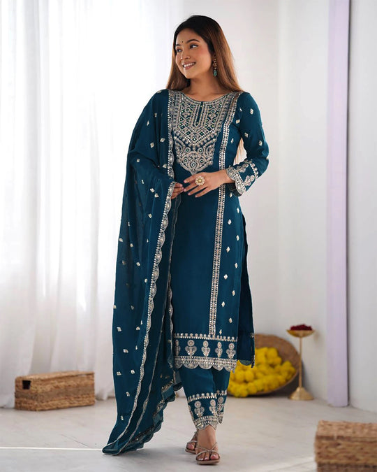 Woman wearing a teal traditional outfit with white embroidery in a minimal indoor setting.