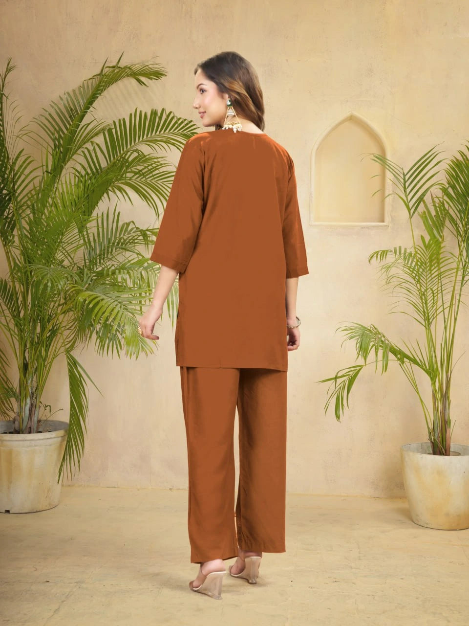 Woman in a brown outfit standing between two potted plants with a beige wall in the background