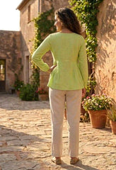 Woman in a green top and beige pants standing in a sunlit outdoor setting with stone walls and plants.